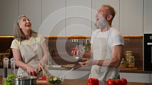 Happy Caucasian senior old couple having fun cooking at home funny man fooling around juggling tomatoes smiling woman