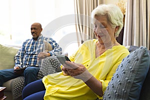 Happy caucasian senior woman sitting on sofa, using smartphone and smiling in sunny living room