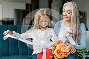 Happy caucasian small granddaughter opens box with gift from elderly grandma with bouquet of flowers