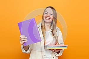 Happy caucasian student girl shows one book to the camera and holds a lot of books isolated over orange studio