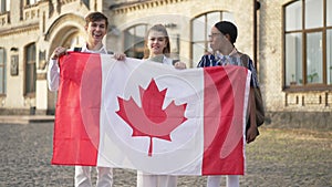Happy Caucasian students posing with African American groupmate holding Canadian flag smiling looking at camera. Three