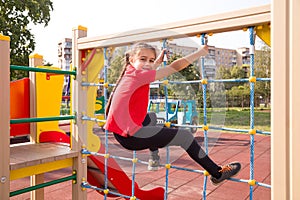 Happy caucasian teenage girl wearing a red T-shirt, having fun on playground, climbing the rope net