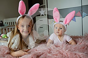 happy caucasian toddler girl 7 years old and her baby sister 6 months old wearing bunny ears headband, lying on bed at