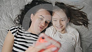 Happy Caucasian woman enjoying family time with daughter lying on bed smiling taking selfies with her smartphone, Social