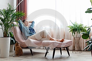 Happy Caucasian woman relax sit in pink chair in cozy home interior with red cat. Smiling young woman have a rest in
