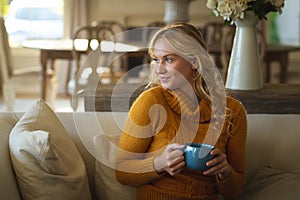 Happy caucasian woman sitting on couch in living room holding coffee cup, smiling and looking away