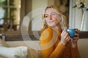 Happy caucasian woman sitting on couch in living room holding coffee cup, smiling and looking away