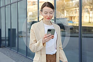 Happy caucasian woman surfing internet on modern smartphone while walking outdoors. Charming female blonde using mobile