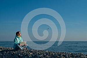Happy caucasian woman working on a laptop while sitting on a pebble beach.