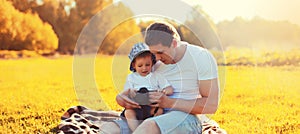 Happy caucasian young father and son child with camera together sitting on the grass in summer sunny evening park