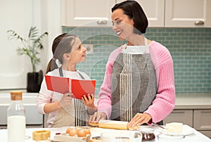 Happy caucasian young mother and little daughter in aprons make cookie dough, use new recipe in book
