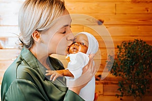 Happy caucasian young woman kissing realistic newborn african american baby doll, indoors.