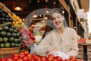 Happy caucasian young woman looks at camera, chooses fresh tomatoes at market.