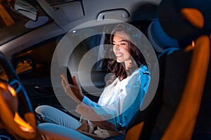 Happy caucasian young woman using smartphone while sitting at passenger front seat of luxury car at night