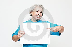 A happy and charming elderly woman holds a blank sheet of paper in her hands and points at it with her finger.