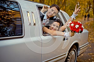 Happy charming newlywed couple is looking through the car window. The bride with the wedding bouquet of red roses is