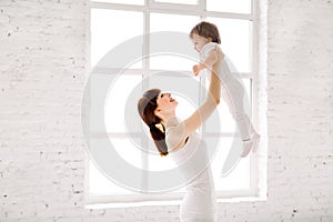 Happy charming sporty mother in white wear holding cute little baby girl up overhead, while standing against big window