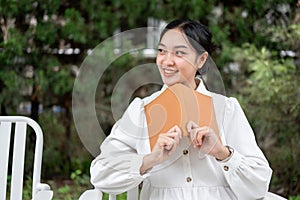 A happy Asian woman sits on a chair with a book in her hand, relaxing in her beautiful green garden