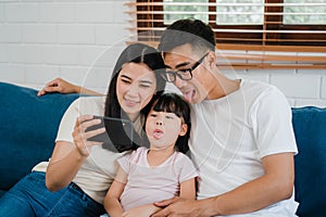 Happy cheerful Asian family dad, mom and kids having fun and using smart phone video call on sofa at house. Self-isolation, stay