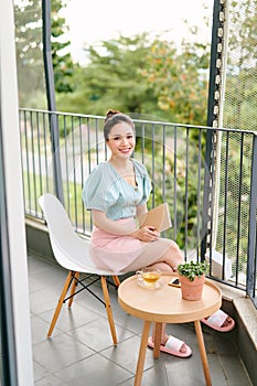 Happy cheerful Asian young woman reading book on the balcony. Relaxation