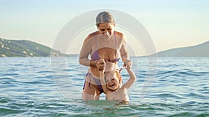 Happy cheerful baby boy with mother enjoying splashing and swimming in the sea. Summertime, holiday, vacation, happiness