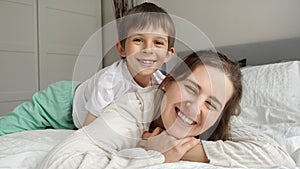 Happy cheerful boy lying with mother in bed and hugging her. Concept of happy family, parents with kids, positive emotions and