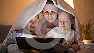 Happy cheerful boys in pajamas showing games on tablet computer to their mother in bed. Family having time together, parenting,