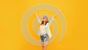 Happy cheerful caucasian smiling young woman raising her hands up wearing shorts, summer straw hat on yellow background