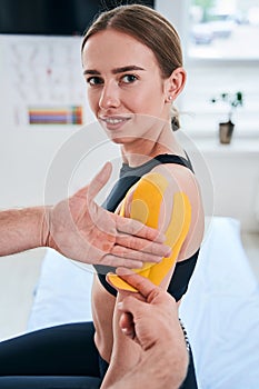 Happy cheerful Caucasian woman getting rehabilitation kinesiology procedure on her shoulder in wellness center