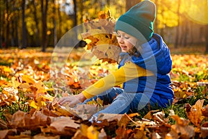 Happy cheerful child, beautiful baby girl in bright colorful warm clothes playing among fallen maple leaves and gathering dry