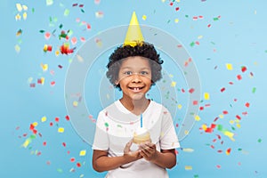 Happy cheerful cute little boy with funny party cone on head holding cupcake and smiling while confetti falling