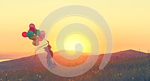 Happy cheerful girl with balloons running across meadow at sunset in summer