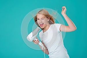 A happy cheerful girl dries her hair with a hair dryer and sings using the hair dryer as a microphone. Blue mint background,