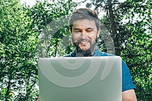 Happy cheerful hipster man with a laptop sitting outdoors on green grass