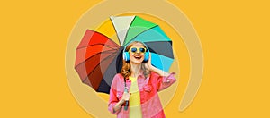 Happy cheerful laughing young woman with colorful umbrella listening to music in headphones wearing pink jacket on yellow studio