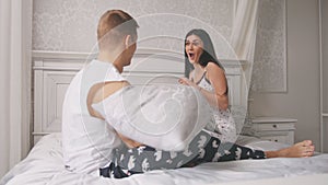 Happy cheerful loving couple having a pillow fight in bed, young attractive guy and girl sitting in pajamas