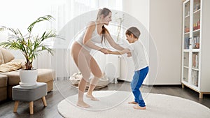 Happy cheerful mother with little son dancing and having fun on carpet in living room