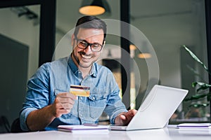 Happy cheerful smiling young adult man doing online shopping or e-shopping satisfied entrepreneur making online payment paying for