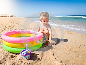 Happy cheerful toodler boy digigng sand on beach and playing with inflatable swimming pool. Child relaxing and having