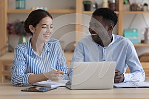 Happy cheerful two diverse employees sharing laptop at workplace