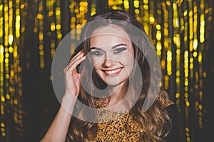 Happy cheerful woman with makeup and long wavy brown hair smiling on golden party background