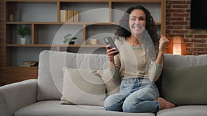 Happy cheerful woman sitting at home couch win with phone cellular device gadget betting good news victory mobile online