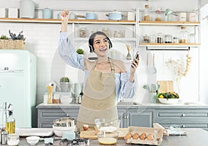 Happy cheerful young asian woman raise smartphone fist good mood wear headphones listen to music in kitchen at home