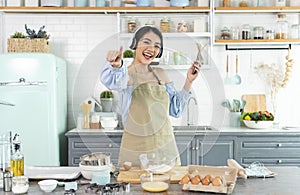 Happy cheerful young asian woman raise smartphone fist good mood wear headphones listen to music in kitchen at home