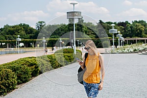 Happy cheerful young blond woman with backpack walking on city street checks her smartphone. Portrait of beautiful 30s