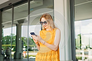 Happy cheerful young blond woman in sunglasses walking on city street checks her smartphone. Portrait of beautiful 30s