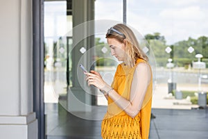 Happy cheerful young blond woman walking on city street checks her smartphone. Portrait of beautiful girl using