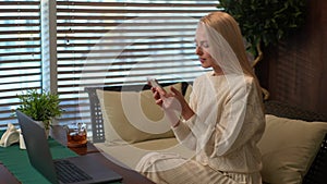 Happy cheerful young blonde woman enjoying using smartphone and social media application with pleasure sitting at table