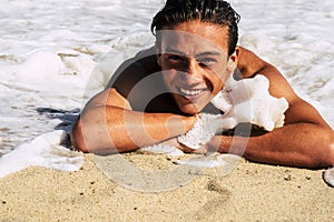 Happy and cheerful young caucasian handsome boy laying on the shore and playgin with wave foam splash smilng and laughing in