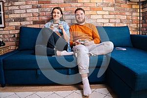 Happy cheerful young couple holding controllers and playing video games on console sitting together